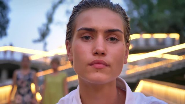 Young modern woman is watching at camera in evening, blurred lights on background