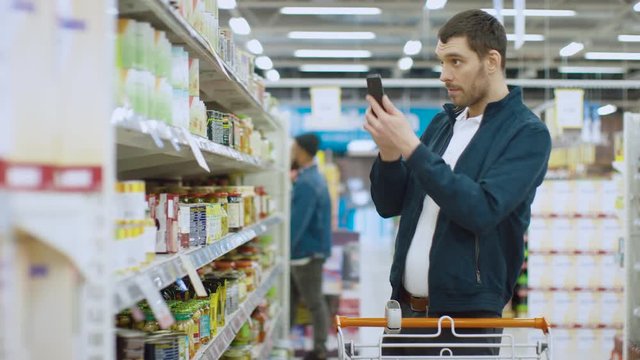 At the Supermarket: Handsome Man Uses Smartphone and Takes Picture of the Can of Goods. He's Standing with Shopping Cart in Canned Goods Section.
