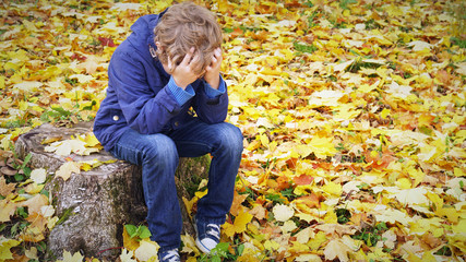 Sad boy sits propping his head with his hands on the stump in autumn park, outdoors