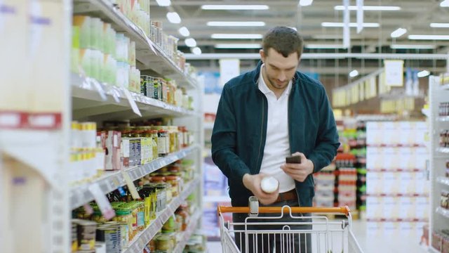 At the Supermarket: Handsome Man Uses Smartphone and Browses Through the Canned Goods Shelf. He's Standing with Shopping Cart in Canned Goods Section.