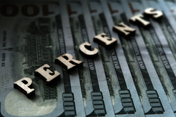 The word PERCENTS is lined with wooden letters on the background of hundred-dollar bills. The concept of financial transactions and bank loans.