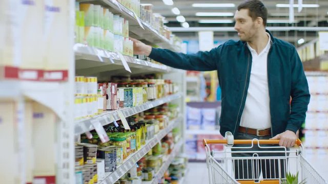 At the Supermarket: Handsome Man Browses Through Shelf with Canned Goods, Chooses Tin Can and Places it into His Shopping Cart.