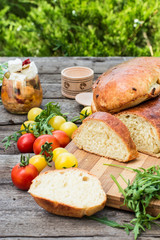 Fresh bread on wooden ground. Yellow tomato and red tomatoes with arugula. Feta cheese with olives and sundried tomatoes. Picnic, dinner outdoor. Still life of food.