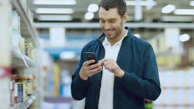 At the Supermarket: Handsome Man Uses Smartphone, Smiles while Standing at the Canned Goods Section. 