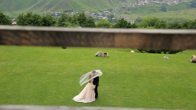 The gentle sensual embrace of the bride and groom under the veil of the bride on their wedding day against the backdrop of green grass and a garden. A view from the side through the fence. Georgia