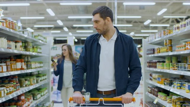 At the Supermarket: Handsome Man with Shopping Cart Walks Through Canned Goods Section, Browsing. Big Store with Lots of Aisles.