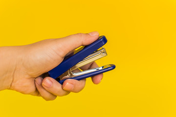 hand holding a blue office stapler on a yellow background - obrazy, fototapety, plakaty