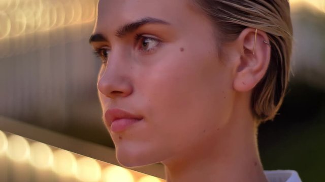 Thoughtful modern woman is watching at camera in evening, blurred lights on background