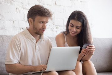 Frustrated husband do online shopping with wife holding credit card dictating bank account details, young spouses making purchases on internet, doubting man look at woman for spending too much