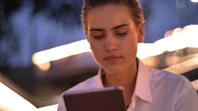 Young modern woman is sitting on ladder, tapping on tablet in evening, blurred lights on background