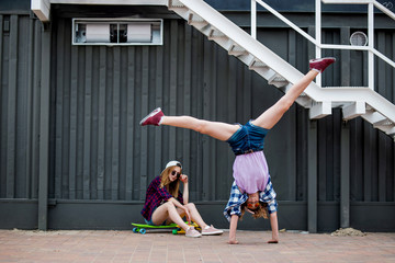 Two pretty blond girls wearing checkered shirts, caps and denim shorts are balancing in front of the black wall on the longboards and having fun while standing upside down. Sport and cool style. © Leika production