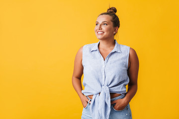 Beautiful smiling girl in shirt and jeans happily looking aside over yellow background isolated. Plus size model