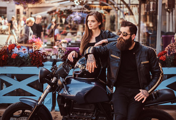 Attractive hipster couple - bearded brutal male in sunglasses dressed in a black leather jacket and his young sensual girl standing near, posing against terrace of cafe.