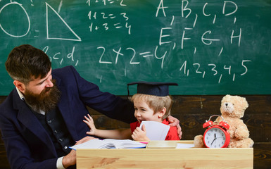 Father with beard, teacher teaches bored son, little boy. Kid fed up with studying, kicks away teacher. Boring studying concept. Teacher and pupil in mortarboard, chalkboard on background.