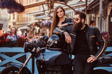Plakat Attractive hipster couple - bearded brutal male in sunglasses dressed in a black leather jacket and his young sensual girl standing near, posing against terrace of cafe.