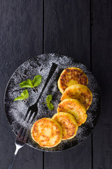 Cottage cheese pancakes on a dark background. Syrniki with fresh mint. Pancakes with cottage cheese on a black plate sprinkled with powdered sugar. Homemade food