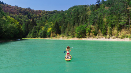 Fototapeta na wymiar une jeune fille à genou et de dos sur son stand up paddle jaune, sur un lac vert entouré d'arbres