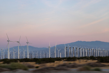 Rows of wind generators set against soft pink, orange sunset sky, mountains in background
