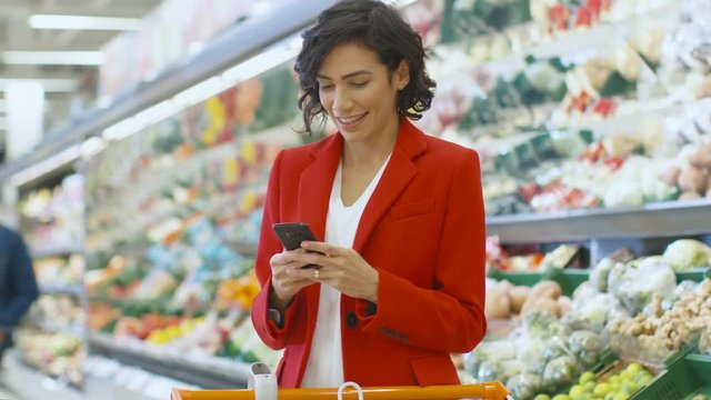 At the Supermarket: Beautiful Woman Uses Smartphone while Standing in the Fresh Produce Section of the Store. Woman Immersed in Internet Surfing on Her Mobile Phone In the Background Colorful Fruits