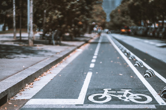 A white bicycle lane sign on a city road asphalt surface in the foreground; safety zone for bikes with dry leaves around, defocused background with strong bokeh, bright autumn day in Barcelona, Spain