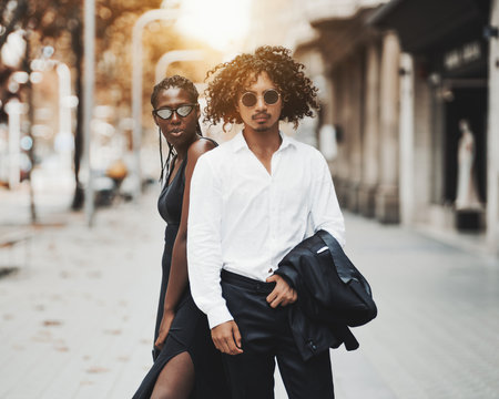 Interracial couple on the city street: a young charming African girl with braids, in a black dress and an asian guy with curly hair, in sunglasses holding a jacket of his suit; sunny autumn day