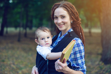 Mom with a baby in a sling reading a book in the Park. Concept of children's education in family...