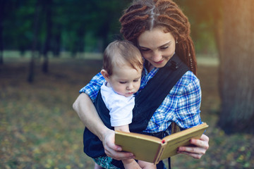 Happy mom with a baby in a sling reading a book in the Park. Children's education in family life