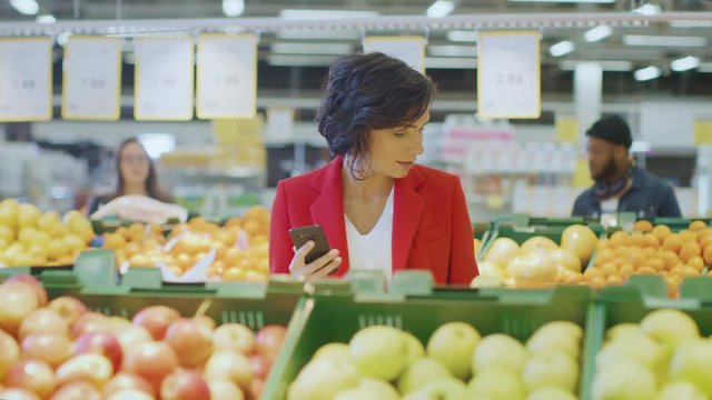At the Supermarket: Portrait of the Beautiful Woman Using Smartphone, Chooses Products In the Fresh Produce Aisle. Woman Immersed in Internet Surfing on Her Mobile Phone