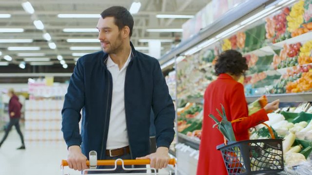 At the Supermarket: Handsome Smiling Man Pushes Shopping Cart, Walks Past Fresh Produce Section of the Store. 