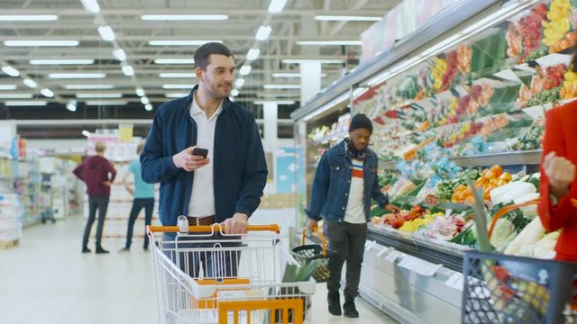 At the Supermarket: Handsome Man with Smartphone, Pushes Shopping Cart, Walks Through Fresh Produce Section of the Store, Chooses Some Products. Other Customers Purchasing Products.
