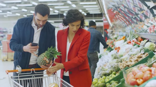 At the Supermarket: Happy Couple Does Shopping, Choosing Fruits and Vegetables in the Fresh Produce Section. Man Uses Smartphone and Pushes Shopping Cart, Woman Places Products into Trolley.
