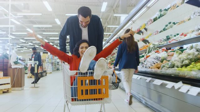At the Supermarket: Man Pushes Shopping Cart with Woman Sitting in it, Happy Couple Has Fun Racing in a Trolley through the Fresh Produce Section of the Store. 