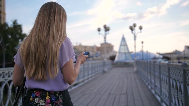 Back view of blonde woman with longe hair walking on city bridge at daytime holding and using cellphone. Beautiful businesswoman in formalwear texting sms on mobile phone while taking a stroll in city