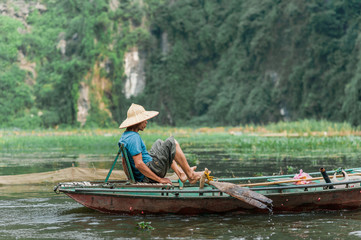 Local man in a rice hat rowing a boat away from the Vung Tram Pier. Traditional paddle-boat trip lets the tourists truly appreciate the serenity and beauty of nature along the Ngo Dong River.