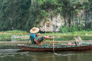 Fototapeta na wymiar Local man in a rice hat rowing a boat away from the Vung Tram Pier. Traditional paddle-boat trip lets the tourists truly appreciate the serenity and beauty of nature along the Ngo Dong River.