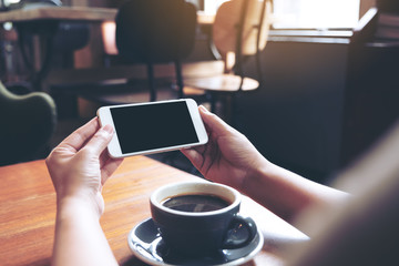 Mockup image of hands holding and using a white mobile phone with blank black screen horizontally for watching with coffee cup on wooden table