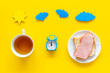 Parts of day. Morning. Time for breakfast. Tea, sandwich near alarm clock, sun and clouds cutout on yellow background top view