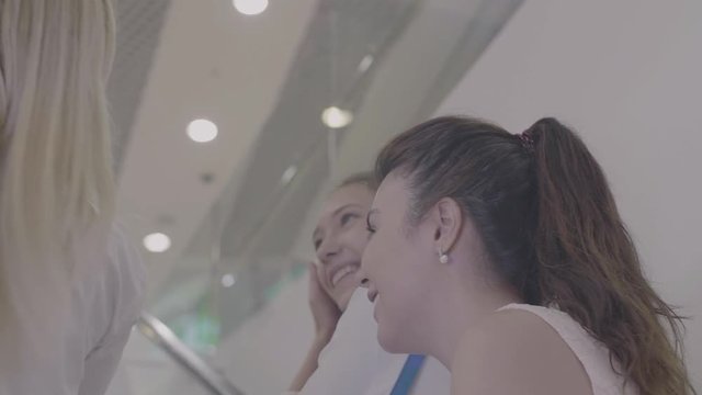 Young Women Going To Work In Business Centre At Escalator, Talking About Something Funny And Laughing. The Moving Stairs Going Up With People. Shopping Mall Or Airport. Trendy Ladies Speaking