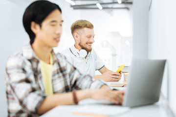 Obraz premium Smiling men. Two young men sitting at the desk in a laconic coworking space and feeling good while using their gadgets