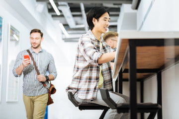 Fototapeta premium Popular coworking space. Enthusiastic young freelancer working on his laptop while a smiling man passing him and looking at the screen of his smartphone