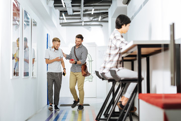 Fototapeta premium Good friends. Cheerful young men walking in a modern coworking space and smiling while looking at the screen of a modern smartphone