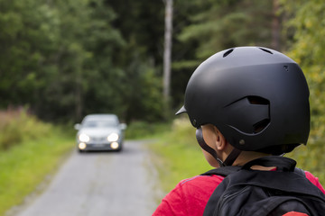 An unidentified little boy looks at the car that is coming in Finland. He has a black bicycle helmet on his head. In the back he has a backpack.