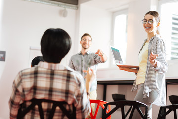 Funny joke. Cheerful friendly young specialist standing with a modern laptop and laughing together with colleagues