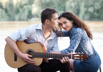 young couple sitting on a log by the river and playing guitar, summer nature, bright sunlight, shadows and green leaves, romantic feelings