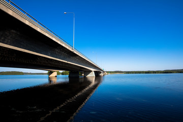 A long concrete bridge, which extends to the horizon in Finland. The bridge crosses the lake. It is noon and the weather is clear and sky is blue. Focal point is the bridge. Image includes a effect.