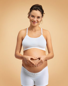 Smiling pregnant woman making a heart gesture with her fingers on beige background. Pregnancy, maternity, preparation and expectation concept