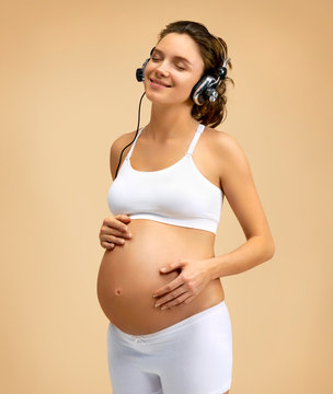 Smiling pregnant woman in headphones touching her belly. Pregnancy, maternity, preparation and expectation concept