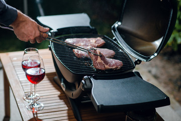 Close up shot of man cooking dinner for two. Beef steak on a grill outdoors on the backyard. Raw steak. BBQ. Date. Glases of wine