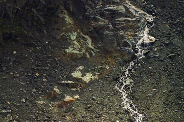 Detailed natural texture of slope from loose stones. Mountain water stream flows down mountainside. Small waterfall on rock. Unusual landscape of majestic nature.