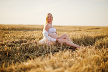 Blonde pregnant woman in wreath field at white underwear clothes on sunset. Happy moments of pregnancy.